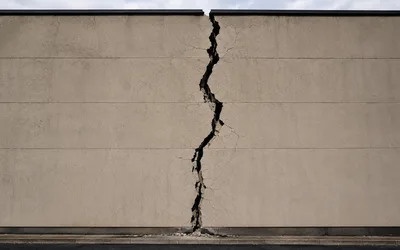 A large smooth concrete wall split by a single deep, jagged vertical crack running from top to bottom, with rubble at its base.