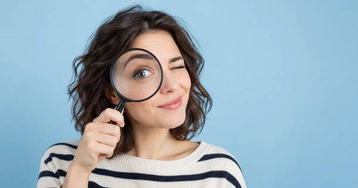 A smiling woman with dark wavy hair in a striped sweater winks while holding a magnifying glass up to one eye against a light blue background