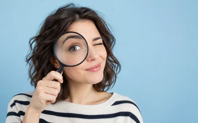 A smiling woman with dark wavy hair in a striped sweater winks while holding a magnifying glass up to one eye against a light blue background