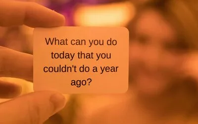 Hand holding a card that reads 'What can you do today that you couldn't do a year ago?' against a warm orange background