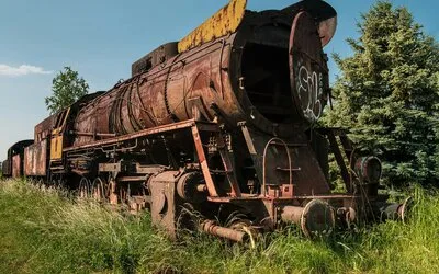 A rusted steam locomotive overtaken by grass and trees, sitting abandoned on overgrown tracks under a clear sky.