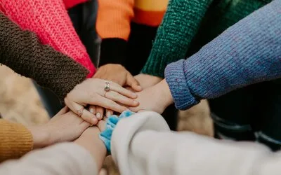 A group of hands stacked together in a circle, each wearing colorful knit sweaters, symbolizing team collaboration and partnership