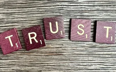 Dark red wooden letter tiles spelling "TRUST" arranged on a weathered gray wood surface