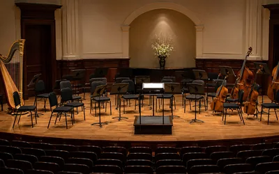 An empty orchestra stage with chairs, music stands, a conductor's podium, a harp, and upright basses arranged and waiting for musicians to arrive.