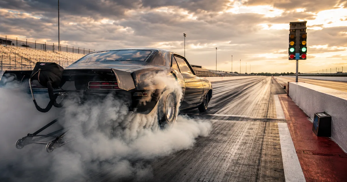 A black muscle car does a stationary burnout at a drag strip start line, rear tires spinning up a thick cloud of smoke while the car stays held in place under green staging lights.