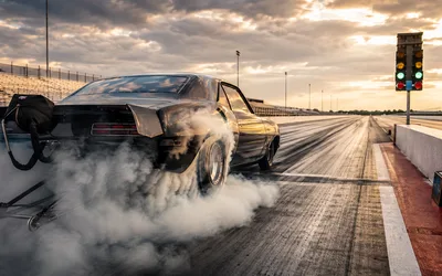 A black muscle car does a stationary burnout at a drag strip start line, rear tires spinning up a thick cloud of smoke while the car stays held in place under green staging lights.