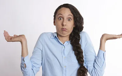 A woman in a blue button-down shirt shrugging with both palms up and a confused expression