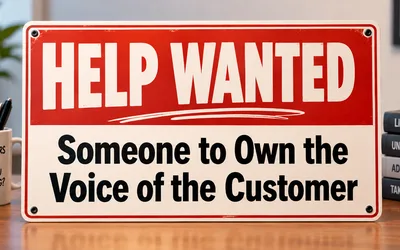 A red and white Help Wanted sign on a wooden desk reading Someone to Own the Voice of the Customer, beside a mug that says Customers talk. Are you listening? and a stack of books labeled Listen, Understand, Advocate, and Take Action