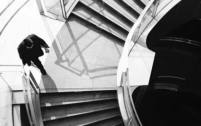 Black and white overhead photograph of a person descending a modern curved staircase with geometric shadows